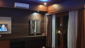 A modern living room with a dark wood panel wall, a large flat-screen TV, a built-in mirror, a sofa, and a sliding glass door with curtains. - Kencana Garden Lembongan (Lembongan Island)