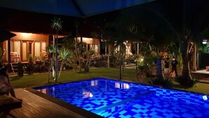 A nighttime view of a pool area with a tiled floor, surrounded by palm trees and a building with warm lighting. - Kencana Garden Lembongan (Lembongan Island)
