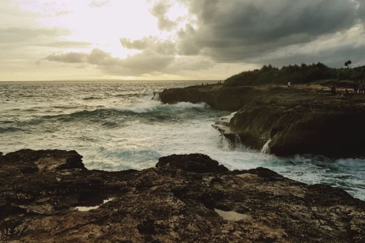 Costa rocosa con olas rompiendo contra las rocas y un acantilado con gente en la cima.