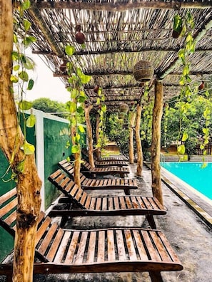 Un espace aménagé au bord de la piscine, avec des chaises longues en bois disposées en rangées sous un toit de chaume, entouré de verdure et d’un mur végétal.