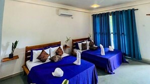 Een hotelkamer met twee bedden, elk voorzien van blauw beddengoed en decoratieve kussens. Er is een airconditioningunit aan de muur en een raam met blauwe gordijnen.