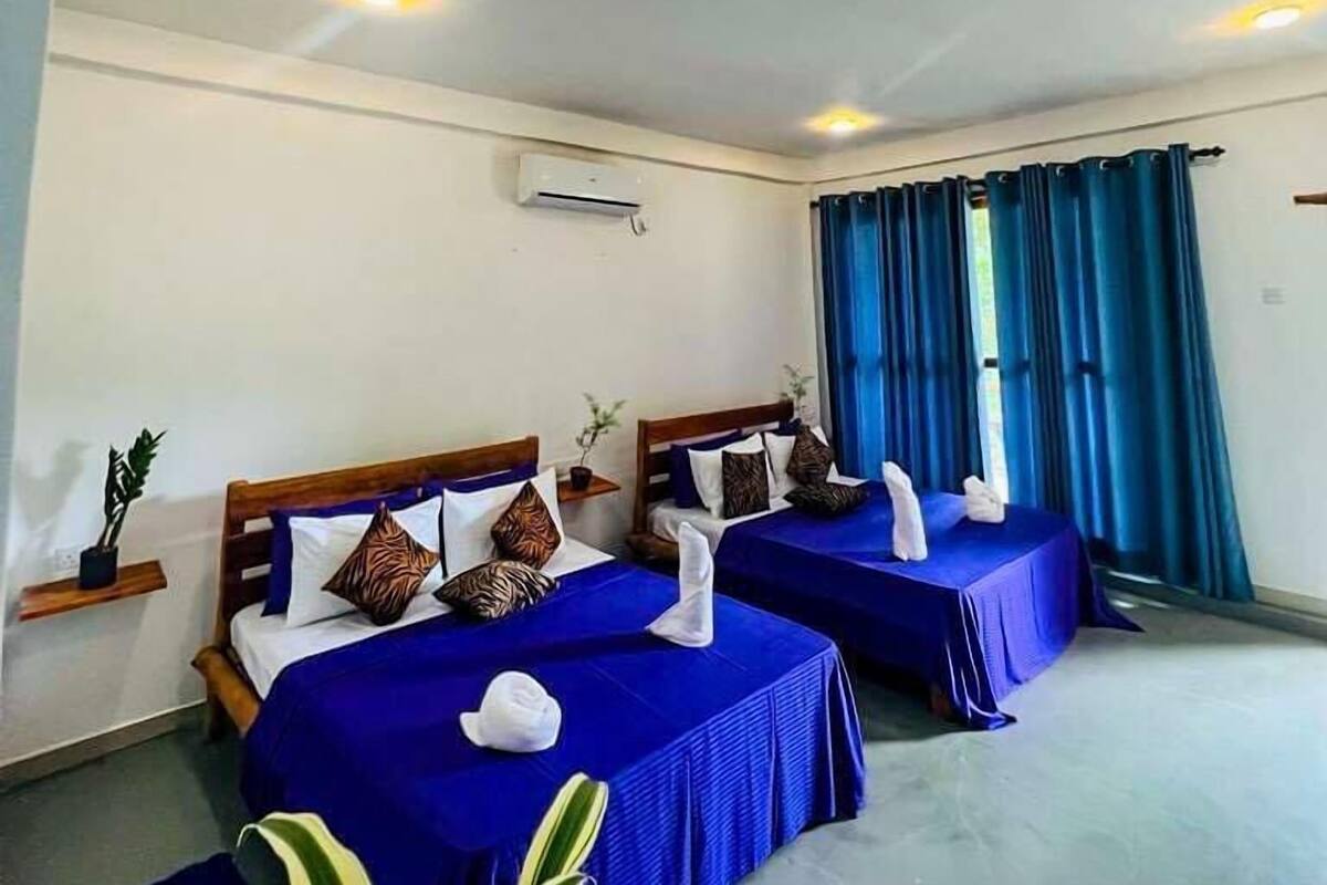 Ein Hotelzimmer mit zwei Betten, jeweils mit blauer Bettwäsche und dekorativen Kissen. An der Wand befindet sich eine Klimaanlage und ein Fenster mit blauen Vorhängen.