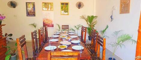 A dining area with a long wooden table set for a meal, surrounded by chairs, with potted plants and wall decorations.