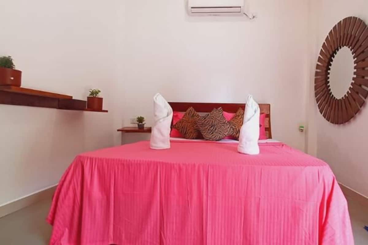 Ein Schlafzimmer mit rosa Bettdecke, einem Holzkopfende, einer Wand-Klimaanlage und einem runden Spiegel.