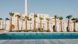 Un espace piscine agrémenté de palmiers et un bâtiment blanc surmonté d’une haute tour.