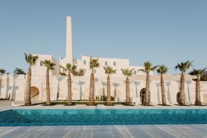 Zona piscina con palme e un edificio bianco con una torre alta.