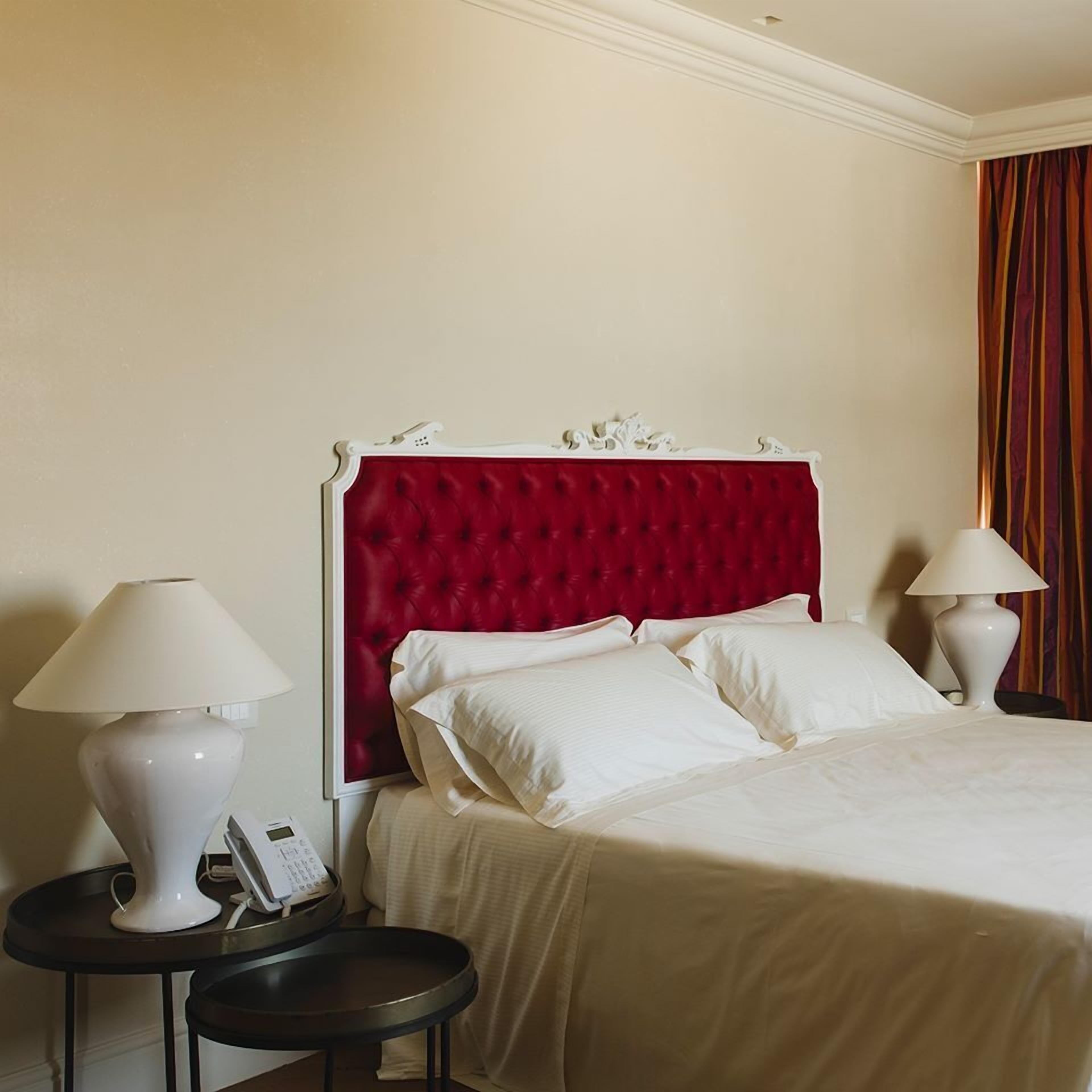 Habitación de hotel con cabecera tapizada en rojo, ropa de cama blanca, dos mesitas de noche con lámparas y un teléfono en una de ellas.