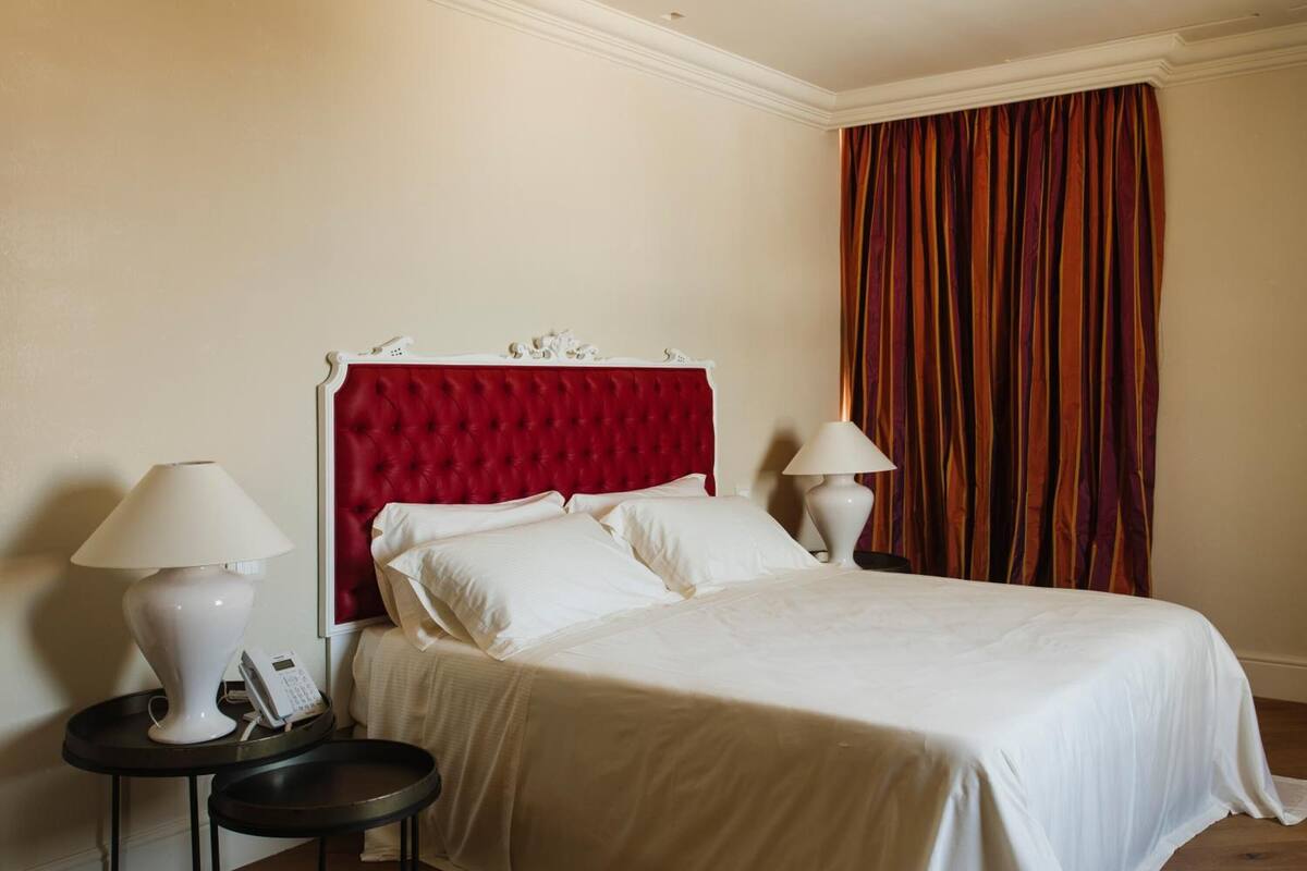 Habitación de hotel con cabecera tapizada en rojo, ropa de cama blanca, dos lámparas de noche y un ventanal con cortinas.