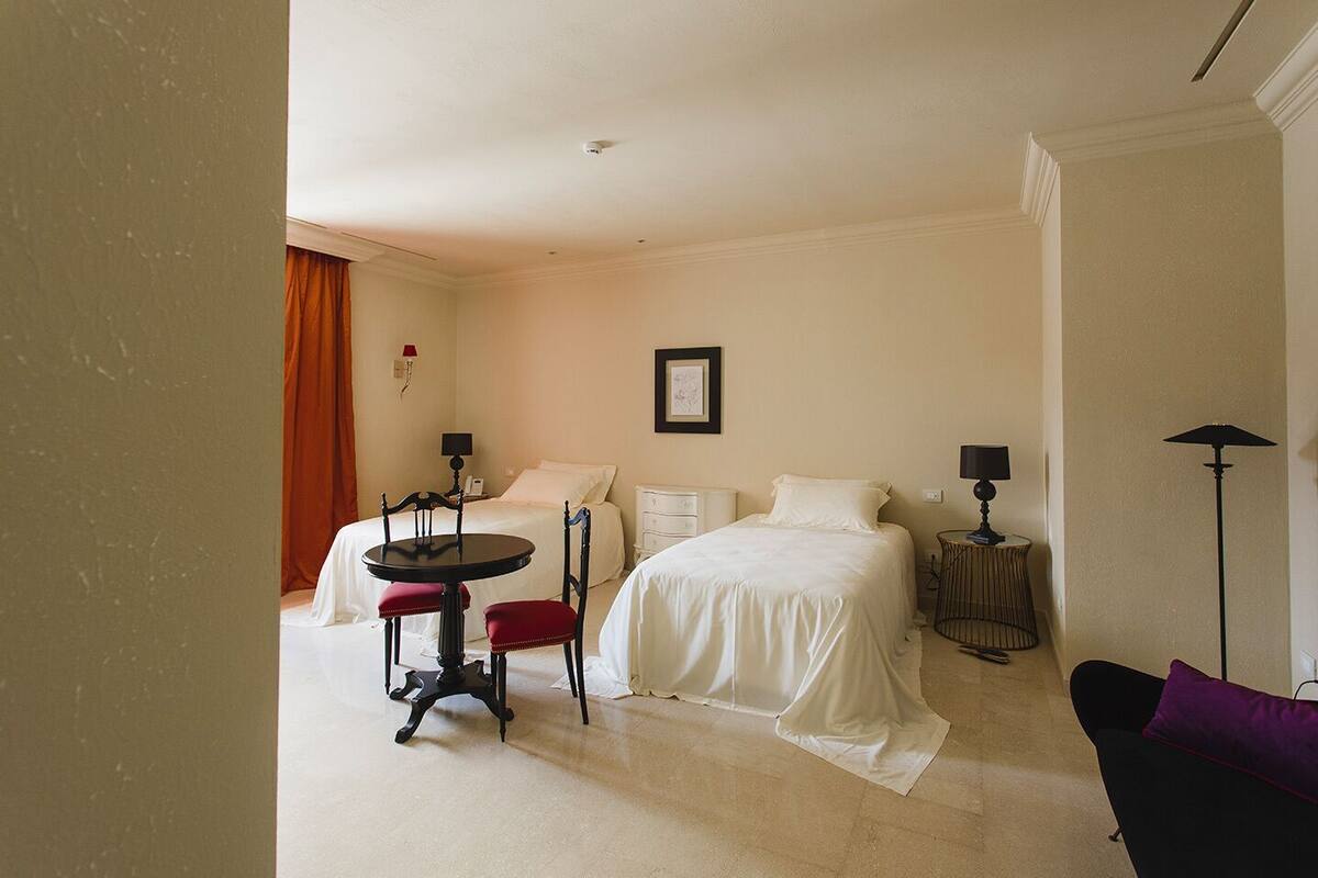 Habitación de hotel con dos camas, una mesa de comedor, una silla, una mesita auxiliar y una lámpara de pie.