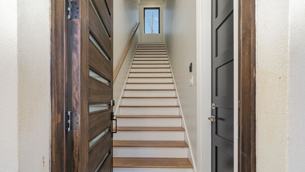 Un estrecho pasillo con una escalera de madera que conduce a una puerta con un ventanal en la parte superior.