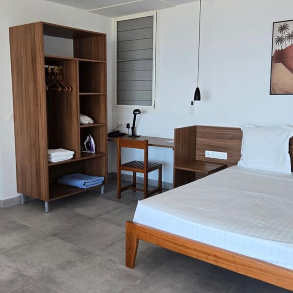 A bedroom with a bed, a wooden wardrobe, a desk with a chair, and a window with blinds.