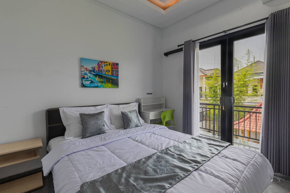 A modern bedroom with a bed, a painting, a desk, and a balcony with a view of buildings.