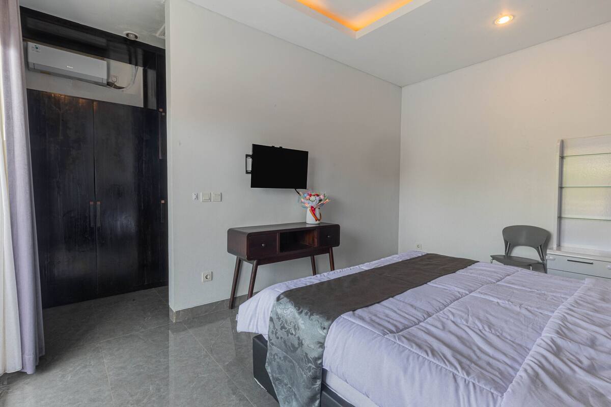 A modern hotel room with a large bed, a flat-screen TV mounted on the wall, a small desk with a vase, and a window with blinds.
