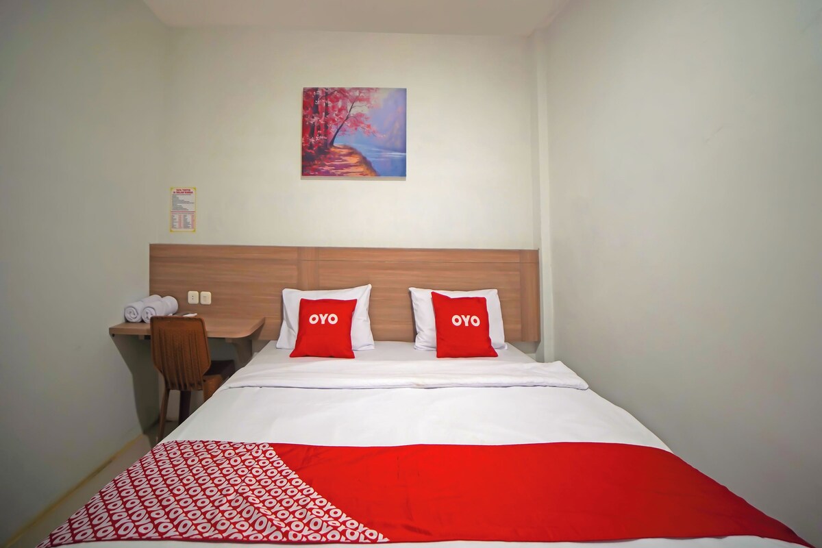 A hotel room with a bed, two pillows with "OYO" branding, a wooden headboard, a painting on the wall, and a small table with towels.