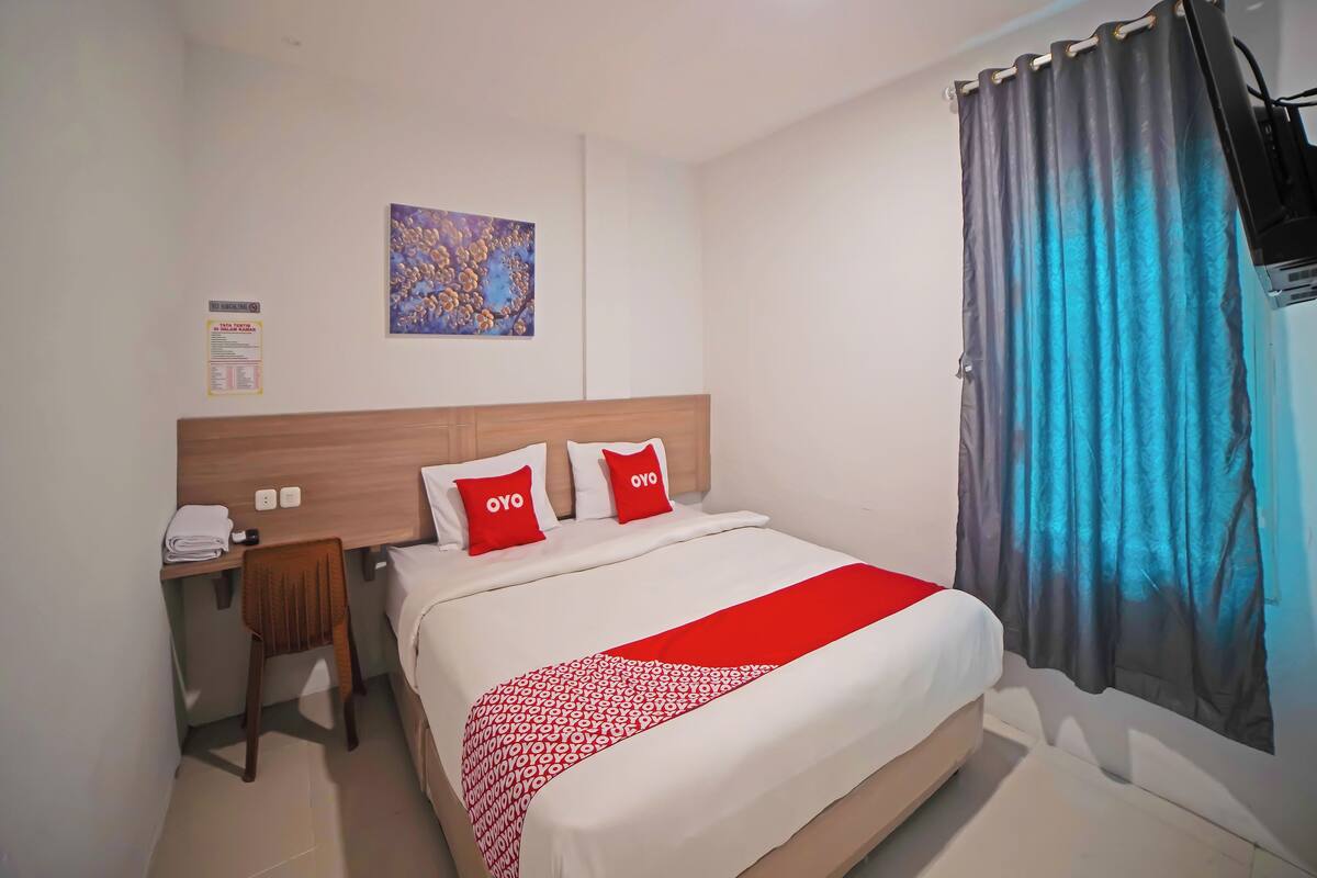 A hotel room with a bed, a desk, a chair, a wall-mounted TV, and a window with curtains.