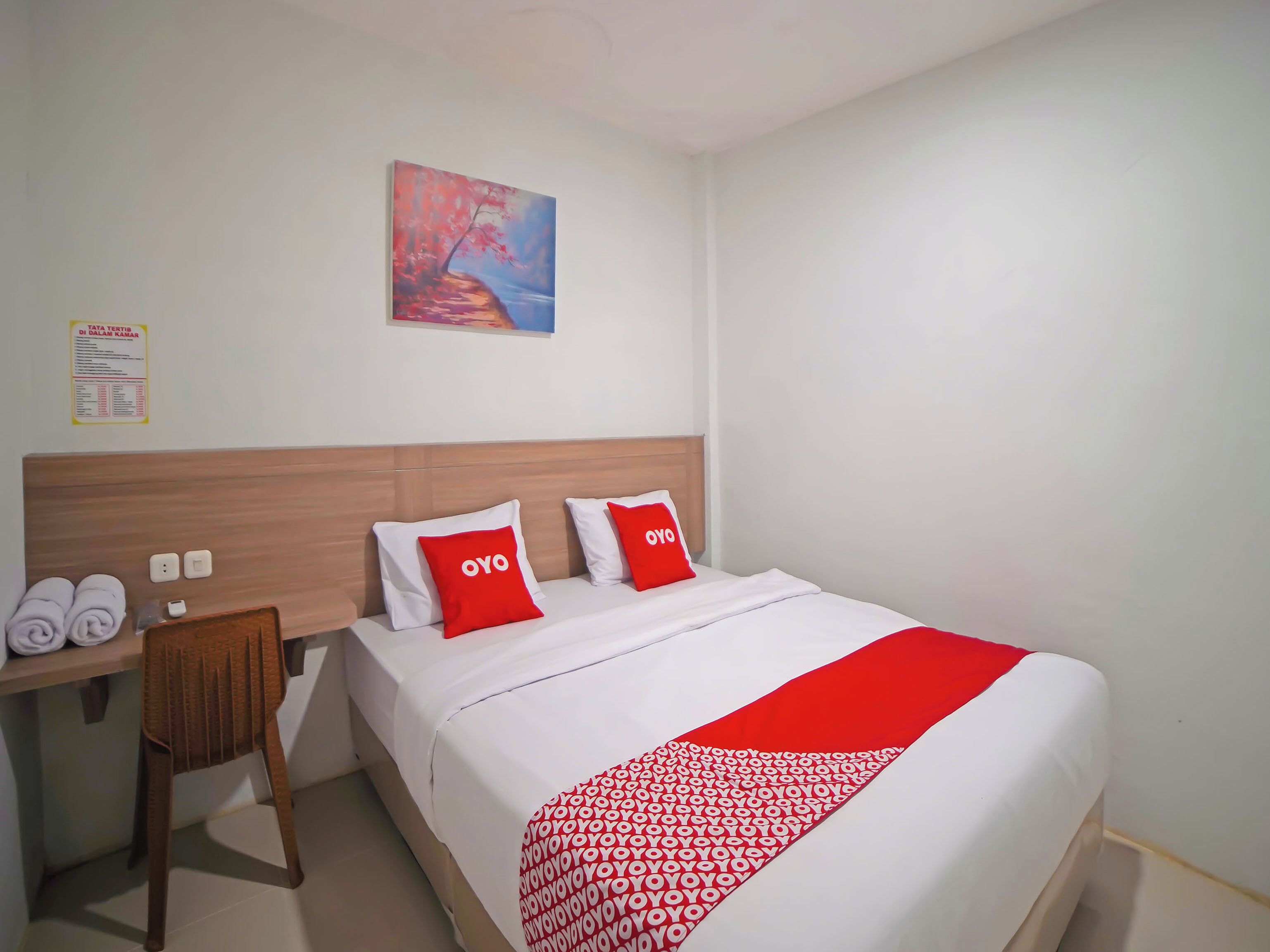 Ein Hotelzimmer mit einem Bett, zwei mit 'OYO' gekennzeichneten Kissen, einem Holzkopfteil, einem Schreibtisch mit Stuhl und einem Bild an der Wand.