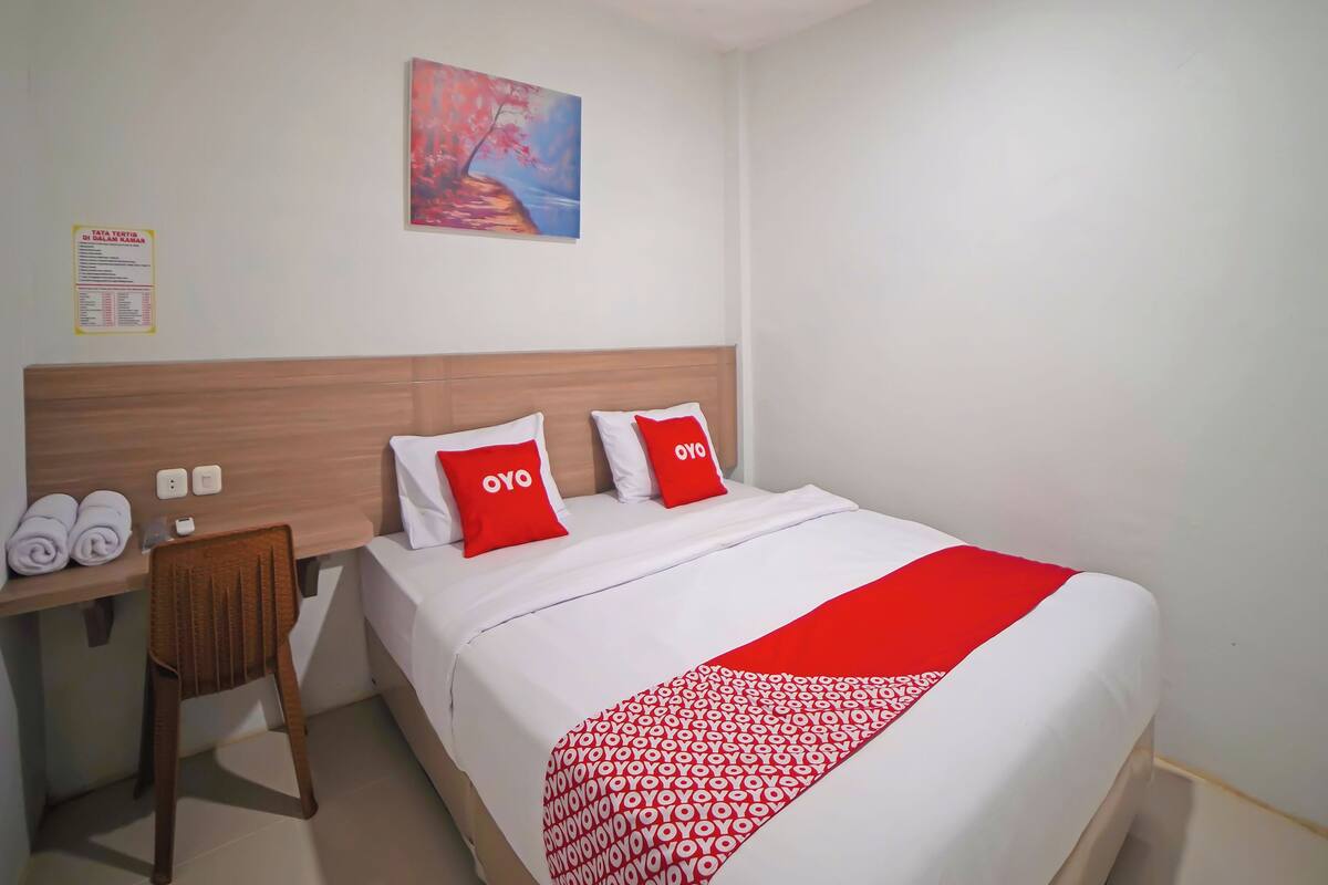 A hotel room with a bed, two pillows with 'OYO' branding, a wooden headboard, a desk with a chair, and a painting on the wall.