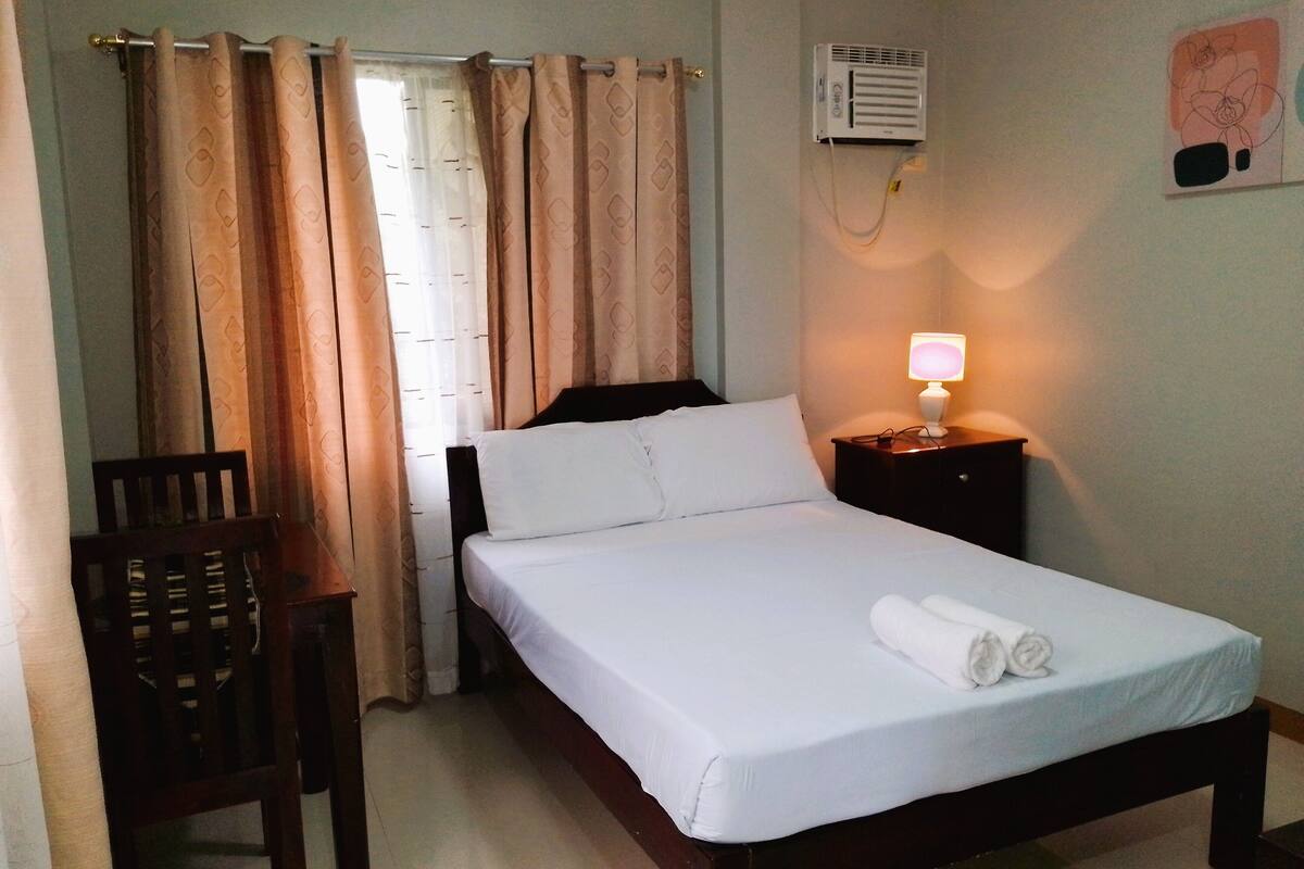 A hotel room with a bed, a chair, a bedside table with a lamp, and an air conditioning unit on the wall.