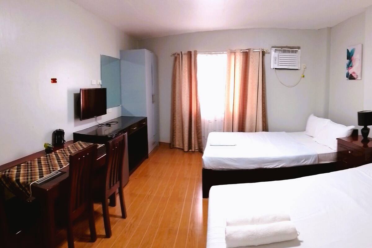 A hotel room with two beds, a desk, a television, and an air conditioning unit.