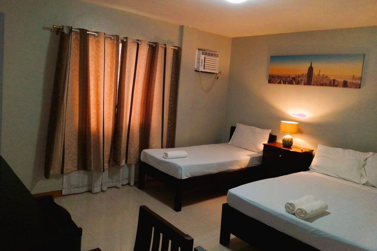 A hotel room with two beds, a nightstand with a lamp, a painting of a cityscape, and a window with curtains.