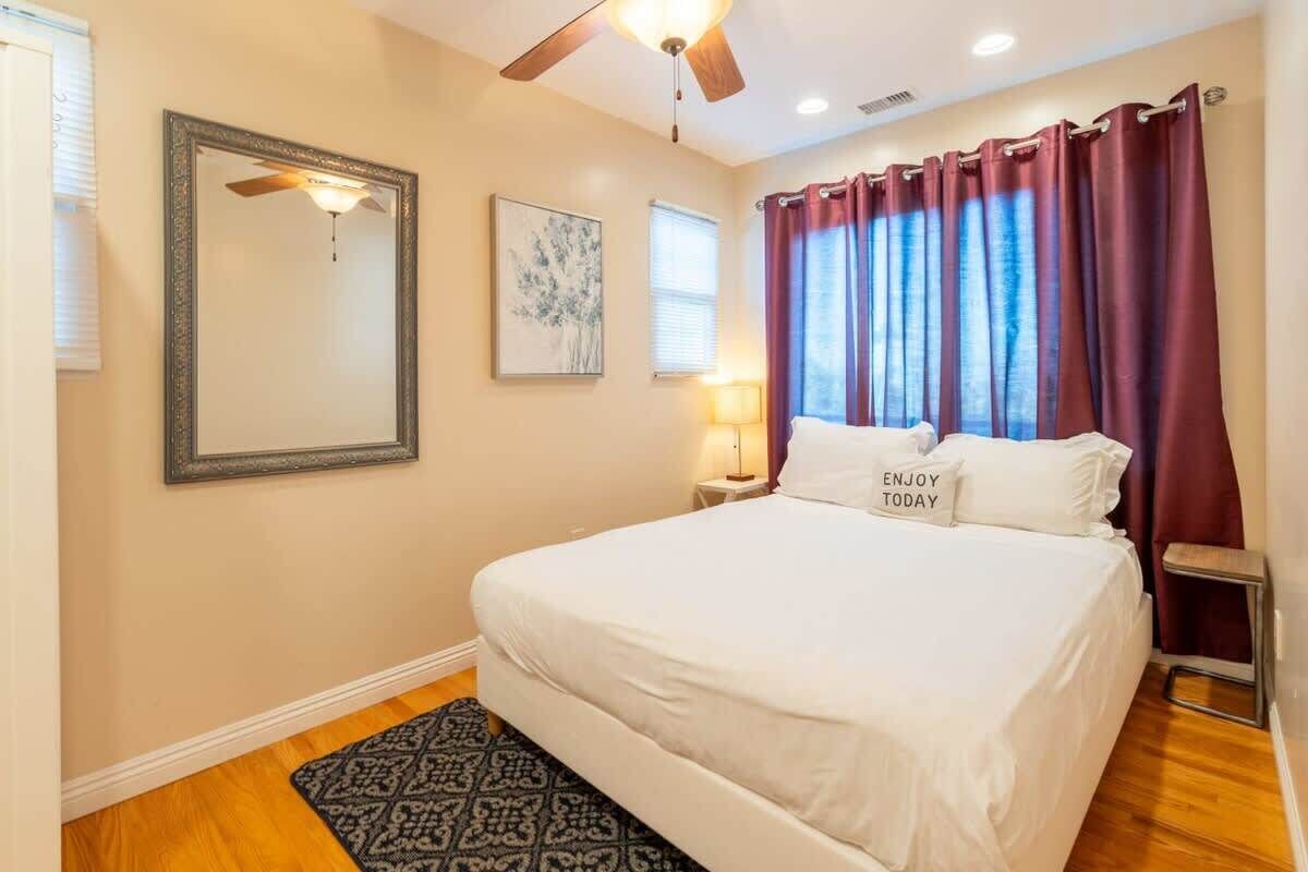 A bedroom with a bed, a ceiling fan, a nightstand with a lamp, a framed picture, and a window with curtains.