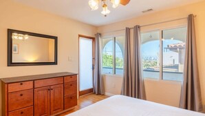 A bedroom with a large bed, wooden dresser, and a large window with curtains. - Serene Point Loma Retreat - Modern 2BR with Views of Point Loma (San Diego)
