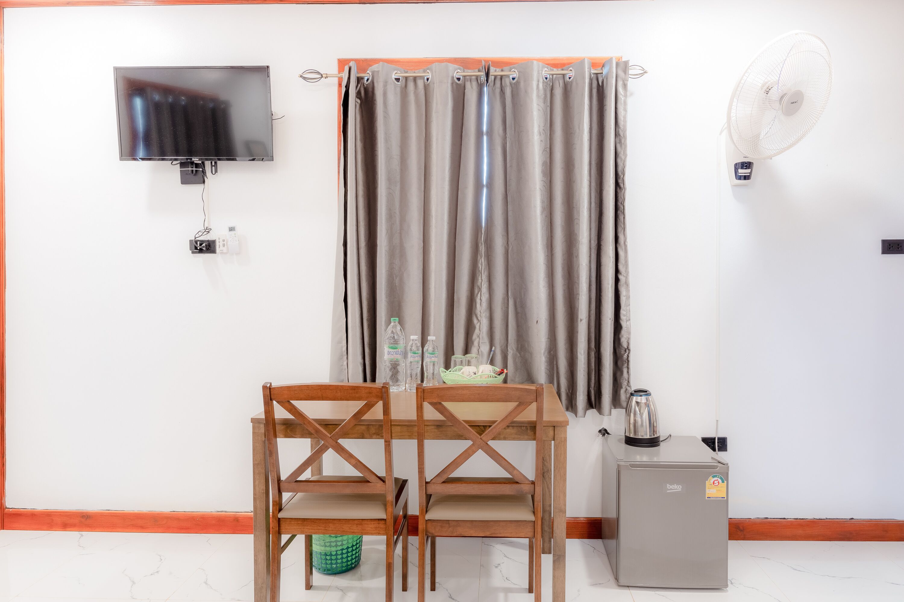 Une pièce comprenant une table en bois, deux chaises, un téléviseur fixé au mur, un ventilateur et un petit réfrigérateur.