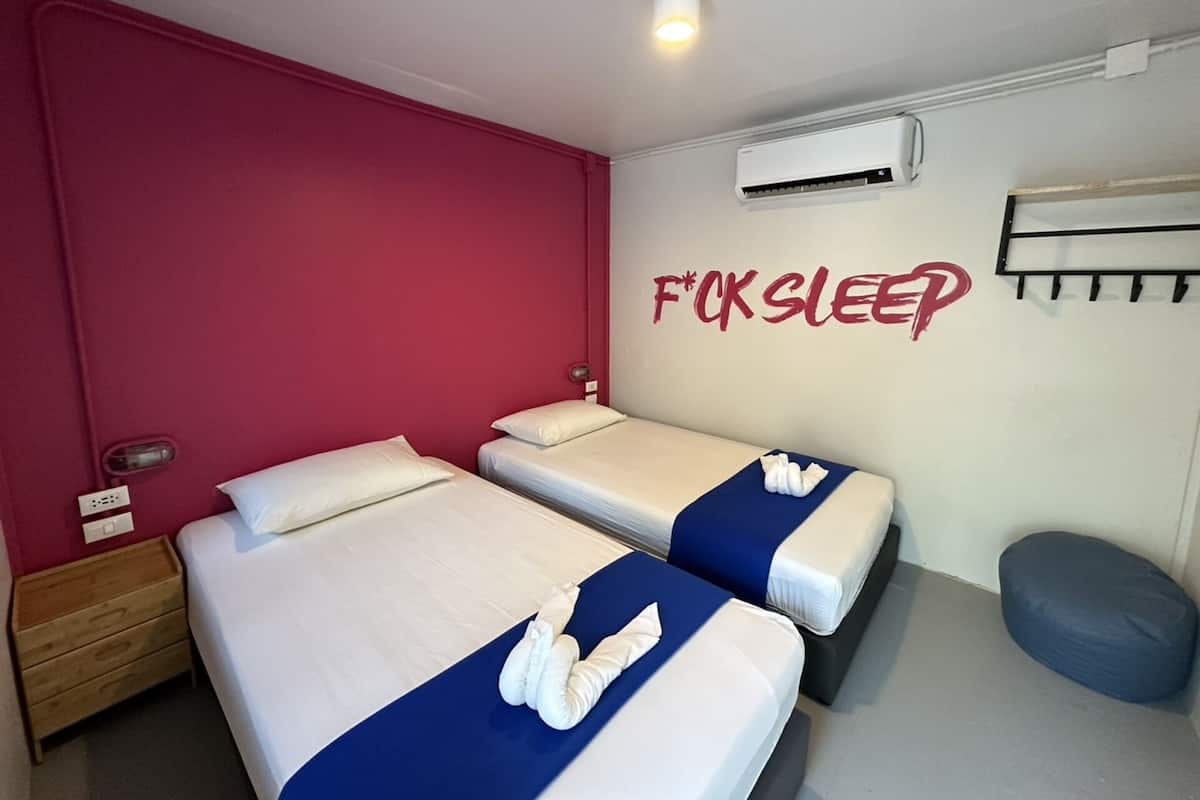 Habitación de hotel con dos camas, una pared roja, un equipo de aire acondicionado y un perchero en la pared.