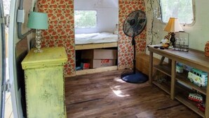 Ein kompaktes Wohnmobil-Interieur mit einem Bett, einem Ventilator, einem kleinen Tisch mit Lampe und einem Regal mit Spielzeug.