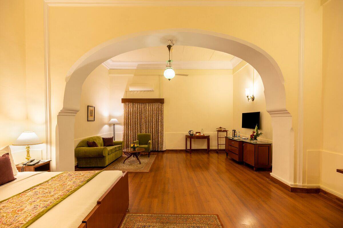 Quarto de hotel com cama, poltrona verde, mesa pequena, televisão e armário de madeira.