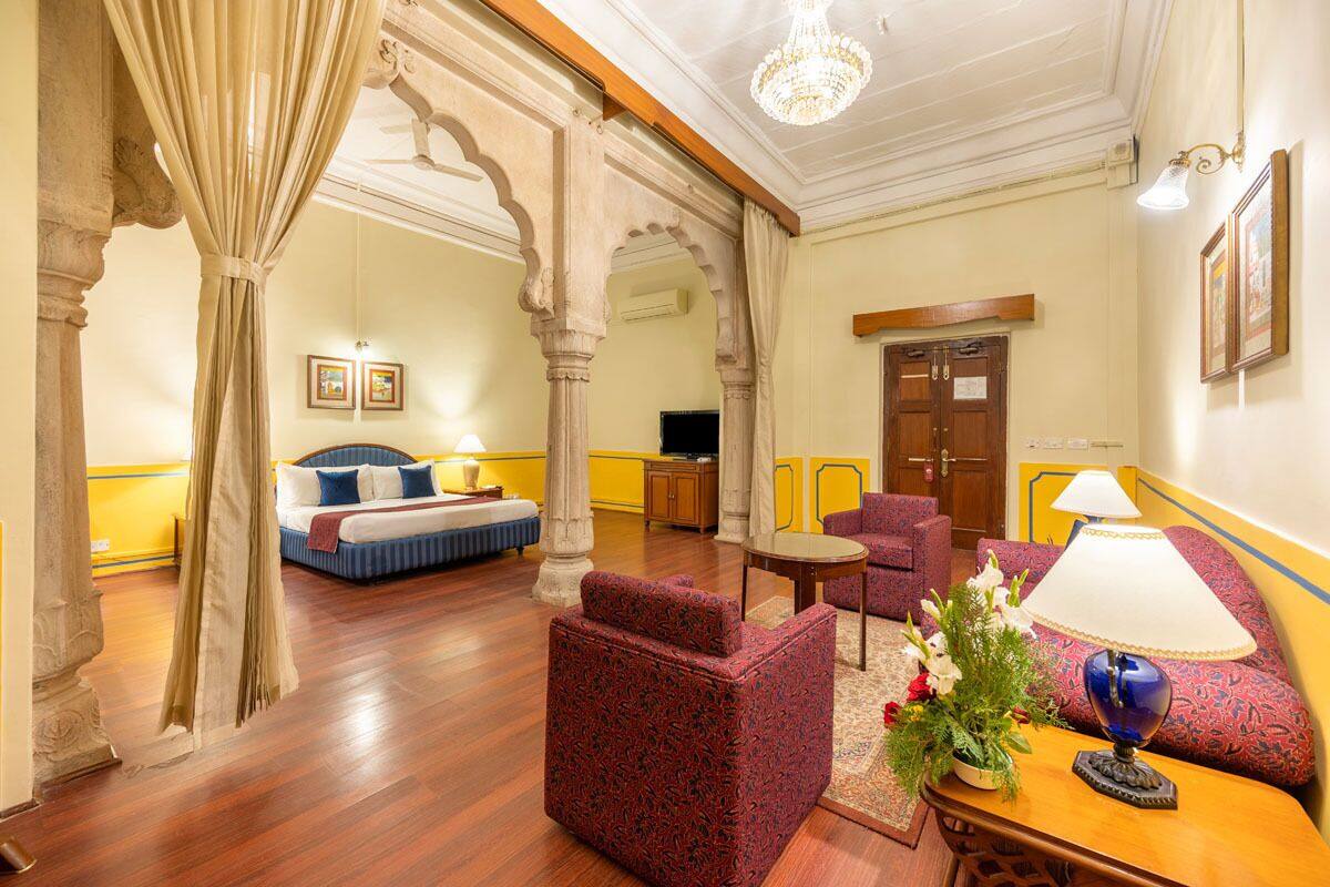 Quarto de hotel com cama, área de estar com sofá e poltrona, pequena mesa com luminária e planta decorativa.
