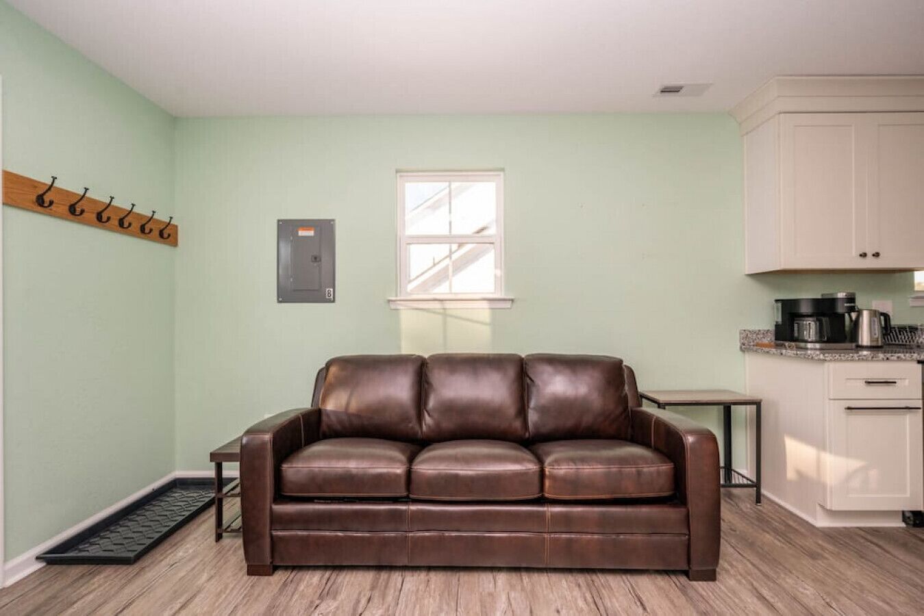 A living room with a brown leather sofa, a small table, a window, and a kitchen area with cabinets and appliances.