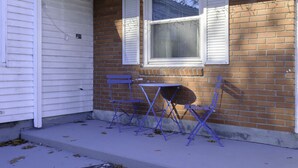 A corner of a house with a brick exterior, a white door, and a window with shutters. There are two blue folding chairs and a small table outside. - The Bradley Studio · Near Mystic, Casinos + USCGA (Groton)