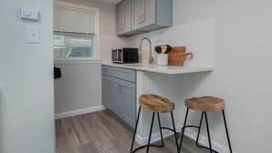 A compact kitchen with grey cabinets, a microwave, a wooden cutting board, and two stools. - The Pratt Studio · Near Mystic, Casinos + USCGA (Groton)