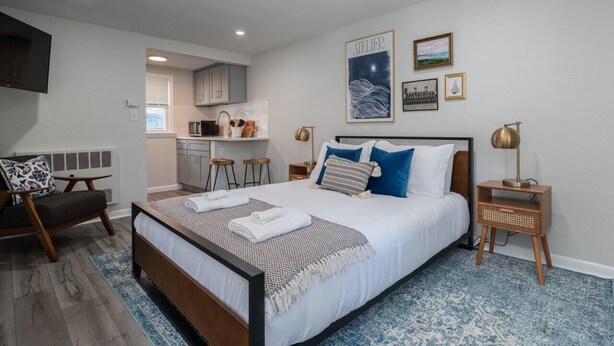 A bedroom with a large bed, bedside tables, a chair, and a view into the kitchen. - The Pratt Studio · Near Mystic, Casinos + USCGA (Groton)