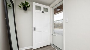 Een witte deur met het nummer 10, een bloempot erboven en een raam met blinds in de aangrenzende muur.