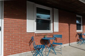 A brick building with white shutters, a small round table, and blue folding chairs outside.