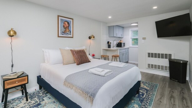 A modern hotel room with a large bed, a desk, a kitchenette, and a TV.