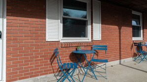 A brick building with white shutters, a small round table, and blue folding chairs outside.