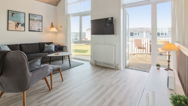 Una sala moderna con un sofá gris, una pequeña mesa de centro y un televisor montado en la pared. Amplias ventanas ofrecen vistas al exterior.