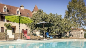 A swimming pool with lounge chairs and umbrellas, surrounded by stone buildings and trees.
