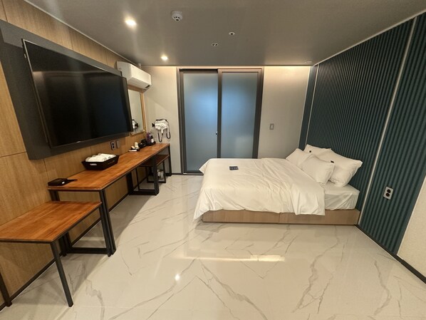 A modern hotel room with a large bed, a desk with a flat-screen TV, a bench, and a wardrobe.