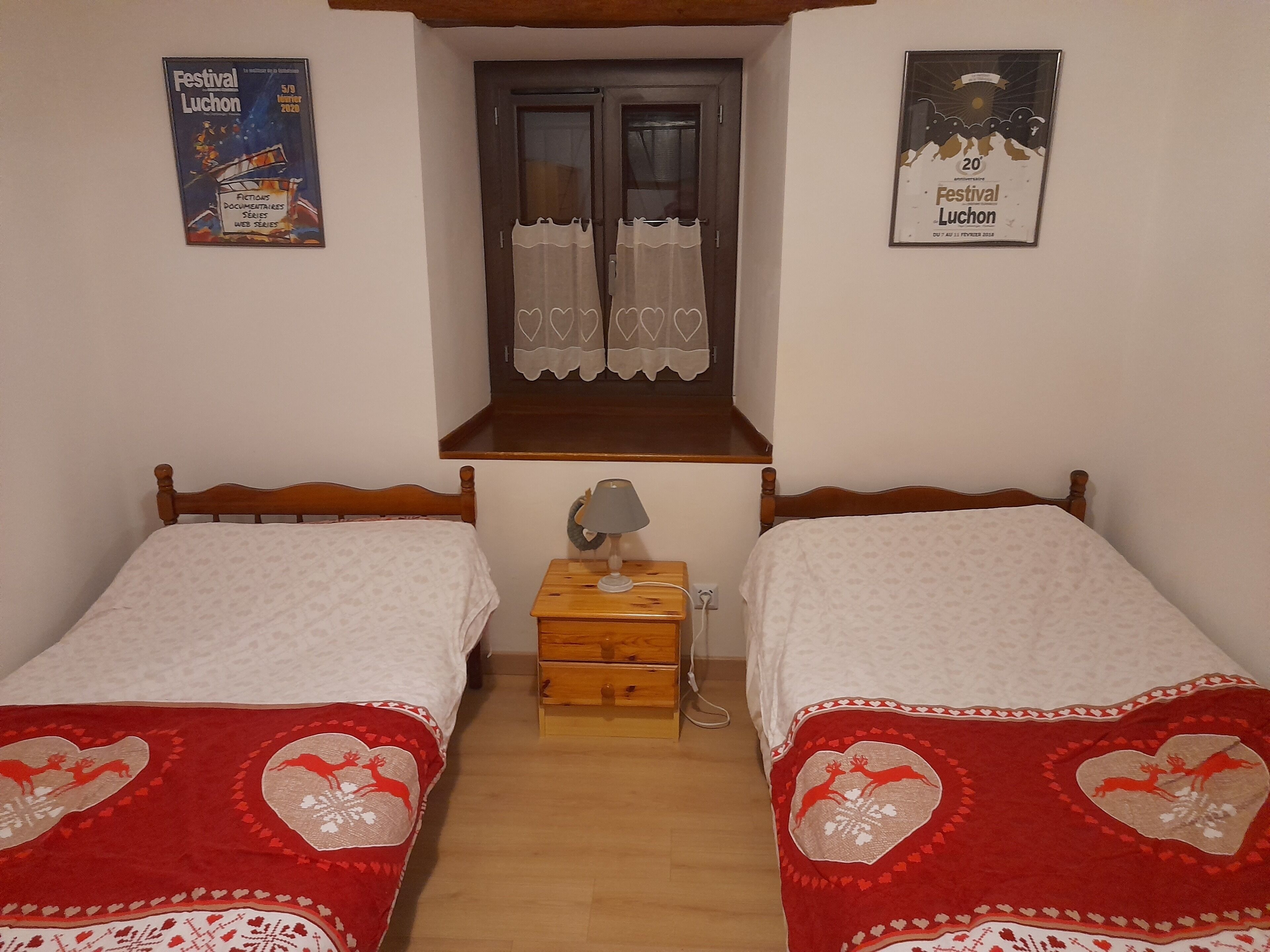 Une chambre avec deux lits, chacun recouvert d’une couverture à motifs de cœurs rouges. Les murs sont ornés de posters, il y a une fenêtre avec des rideaux et une petite table de chevet en bois sur laquelle se trouve une lampe.