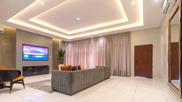 Een moderne woonkamer met een grijze bank, een flatscreen televisie en een groot raam met gordijnen.