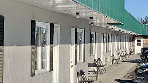 Fila de habitaciones con paredes blancas y contraventanas negras, un toldo verde y un estacionamiento con sillas y un vehículo azul.