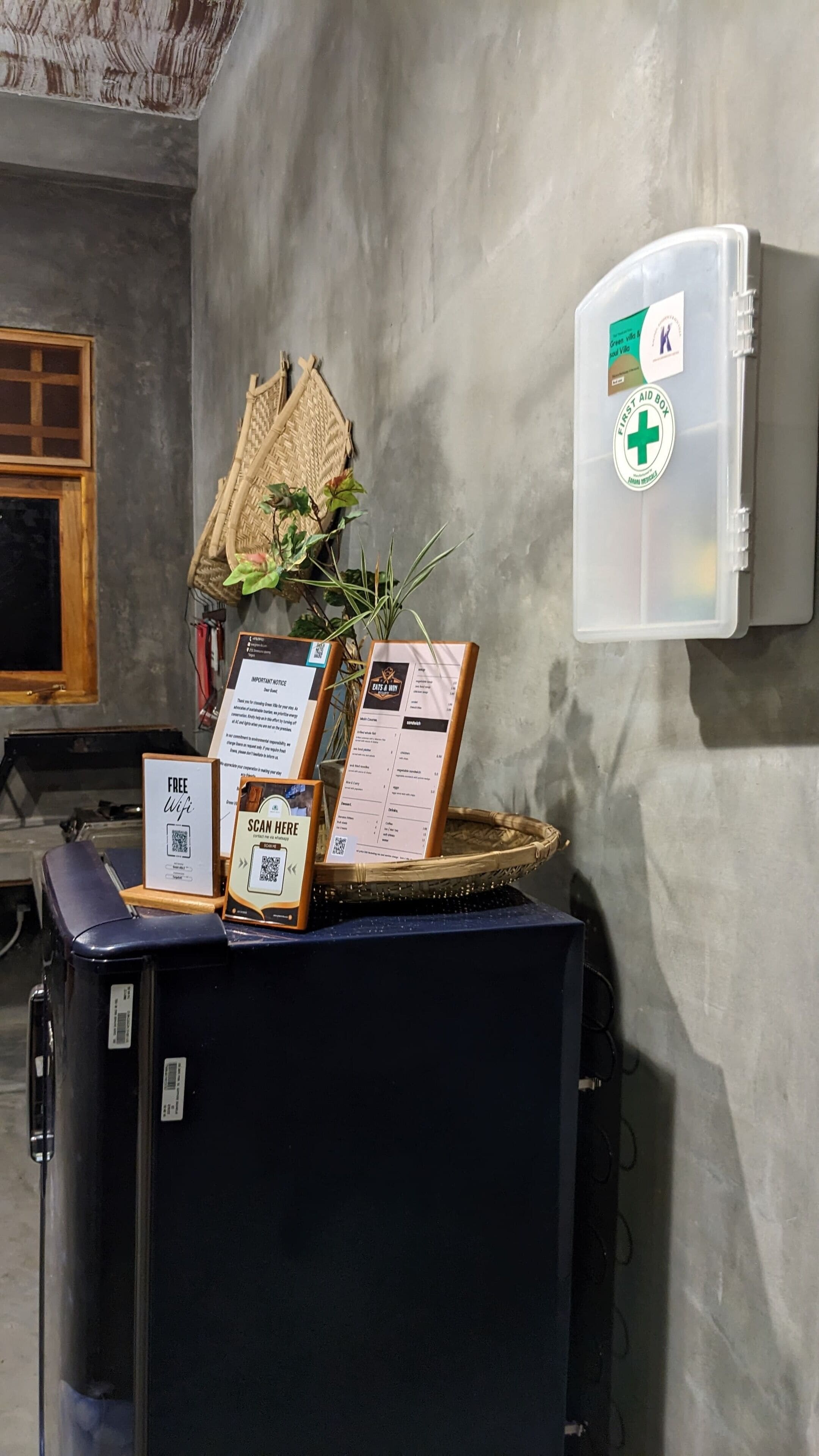 A room with a wall-mounted first aid box, a wicker chair, a plant, and a table with informational signs and a QR code.