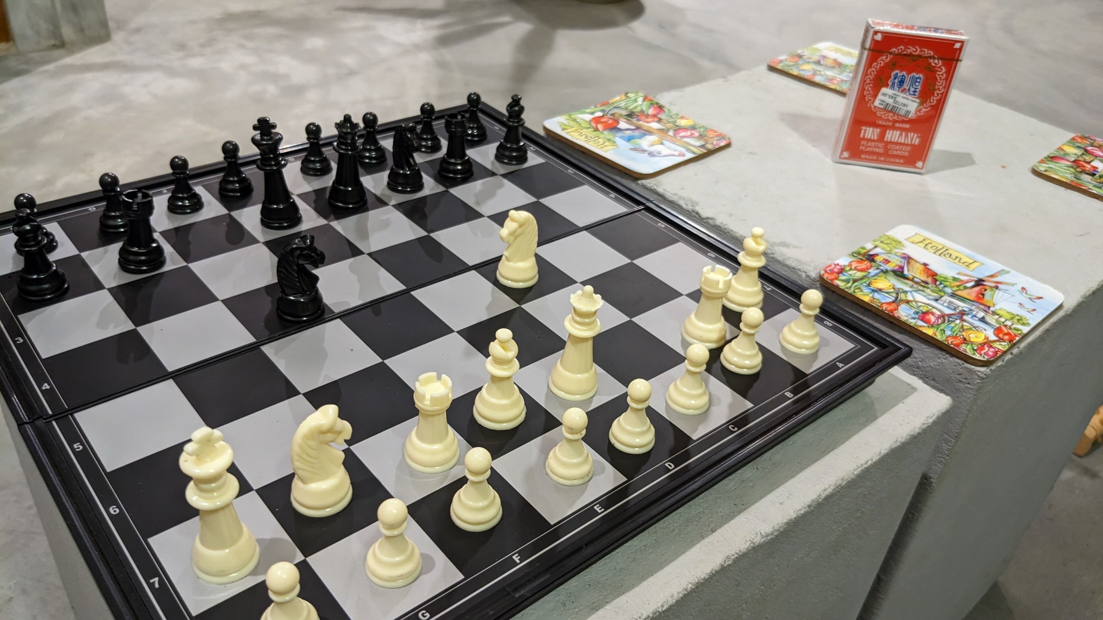 A chessboard with pieces set up for a game, a card game table with cards, and a box of Tic Tac Toe game pieces.