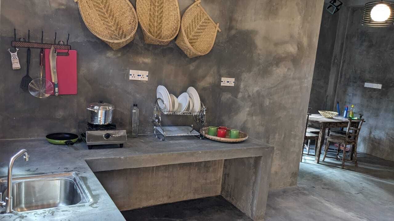 Una cocina con un lavamanos, una estufa y un escurreplatos. Hay cestas colgadas en la pared y una mesa con sillas al fondo.