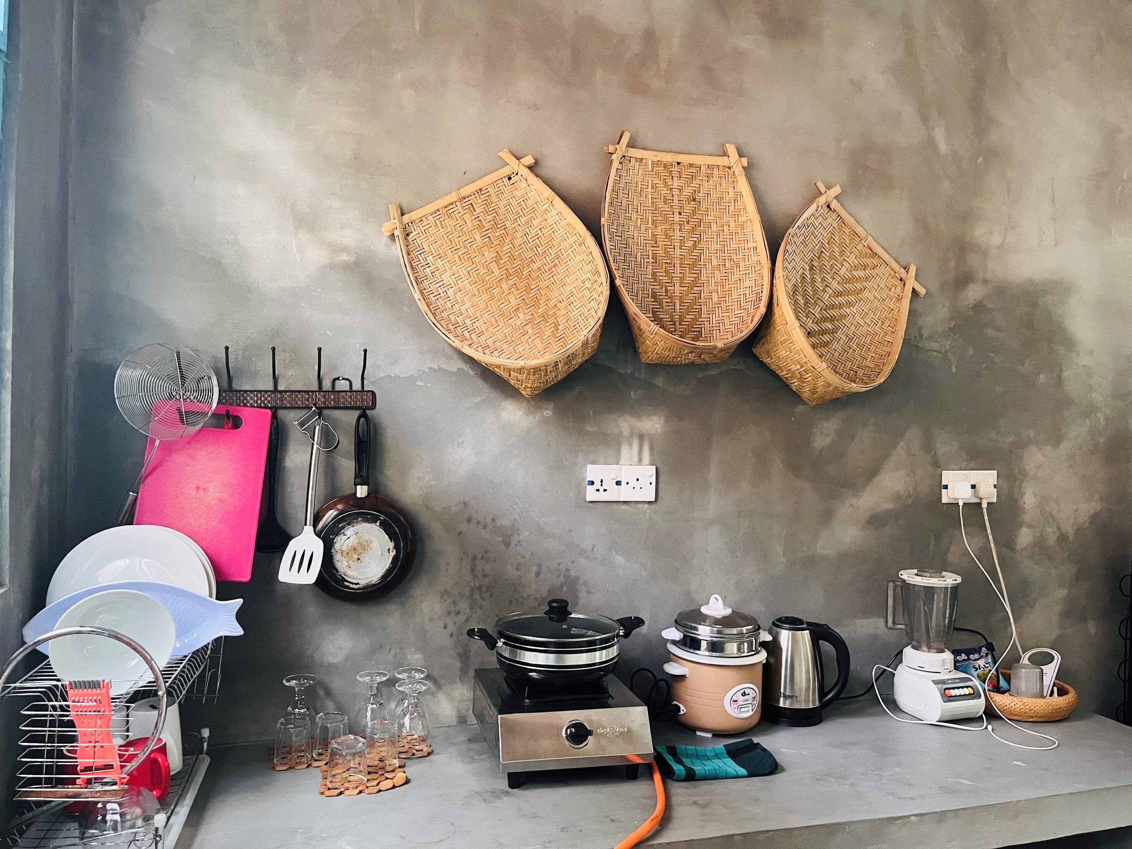 Eine Küche mit Gasherd, Slow Cooker, Wasserkocher und Mixer. An der Wand befinden sich hängende Körbe und ein Geschirrgestell mit Tellern und Gläsern.