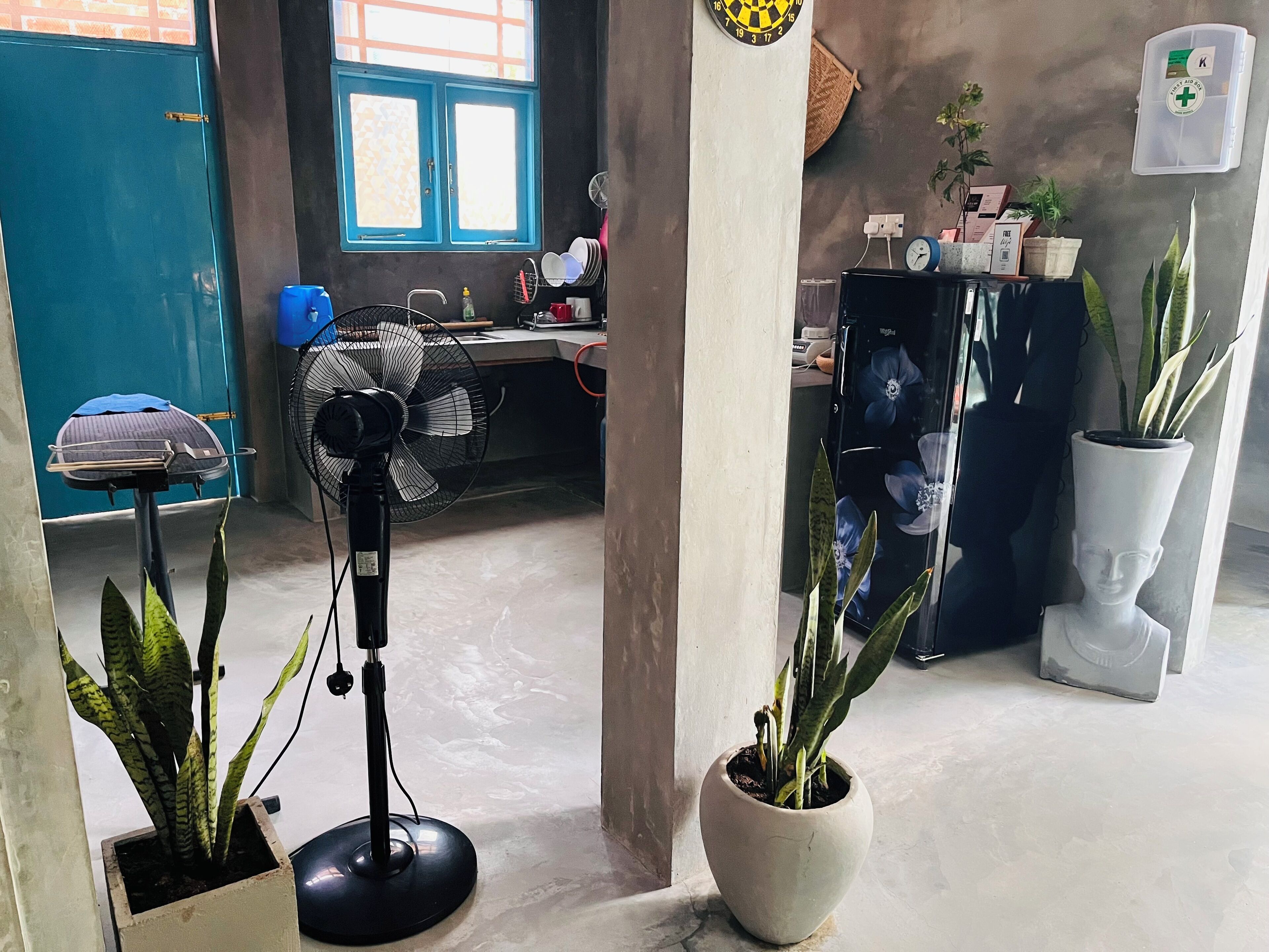 Ein Zimmer mit Schreibtisch, Stuhl, Standventilator und zwei Blumentöpfen.